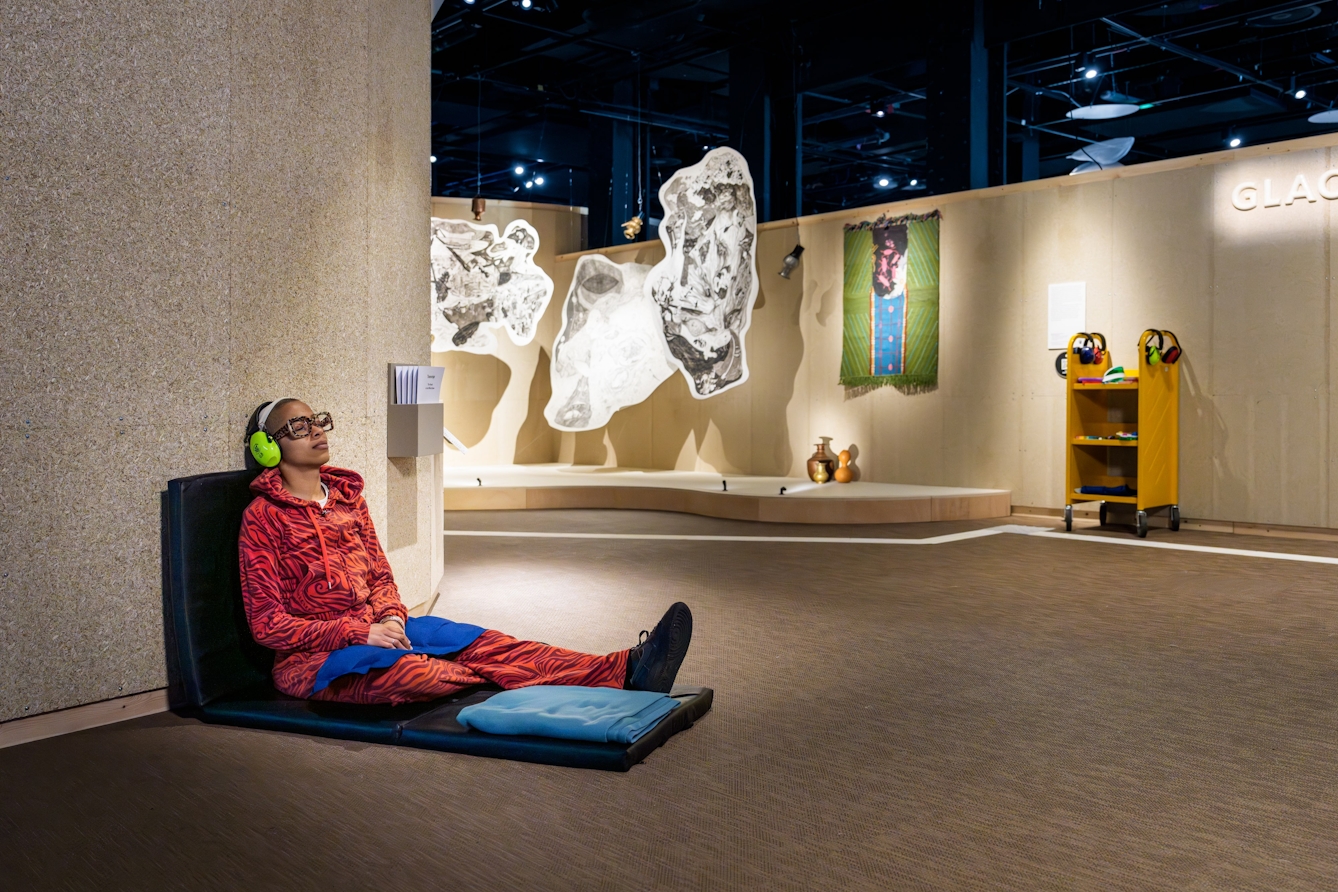 Person wearing ear defenders sitting on a cushioned pad on the floor that is propped up against a wall.