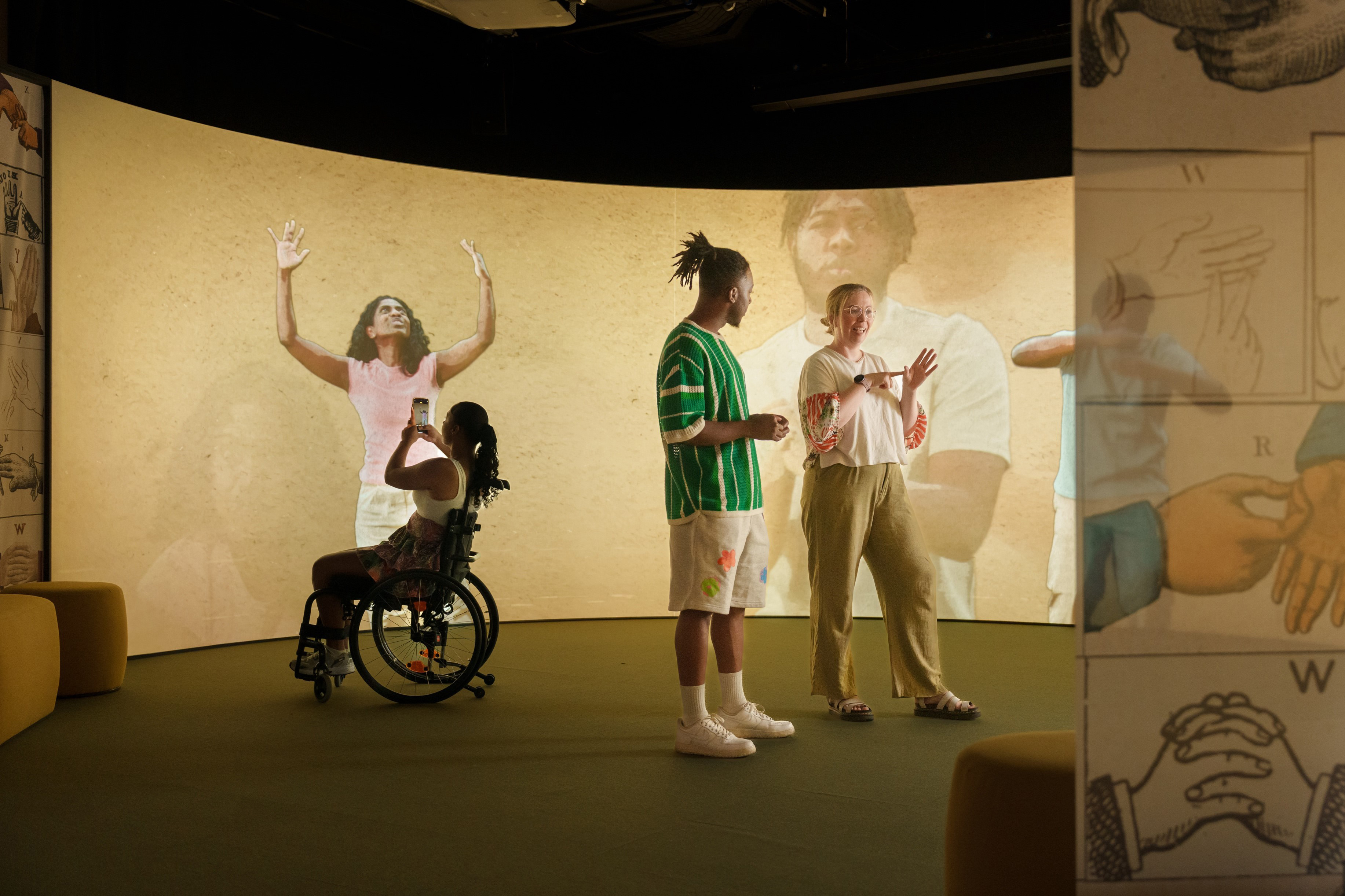 A curved projection screen shows people moving and gesticulating. In front of the screen is a person in a wheelchair taking a picture of the screen with their camera and two other people, standing with their backs to the screen and conversing in sign language. to the right of the image is a transparent curtain printed with historical illustrations of a signing alphabet.