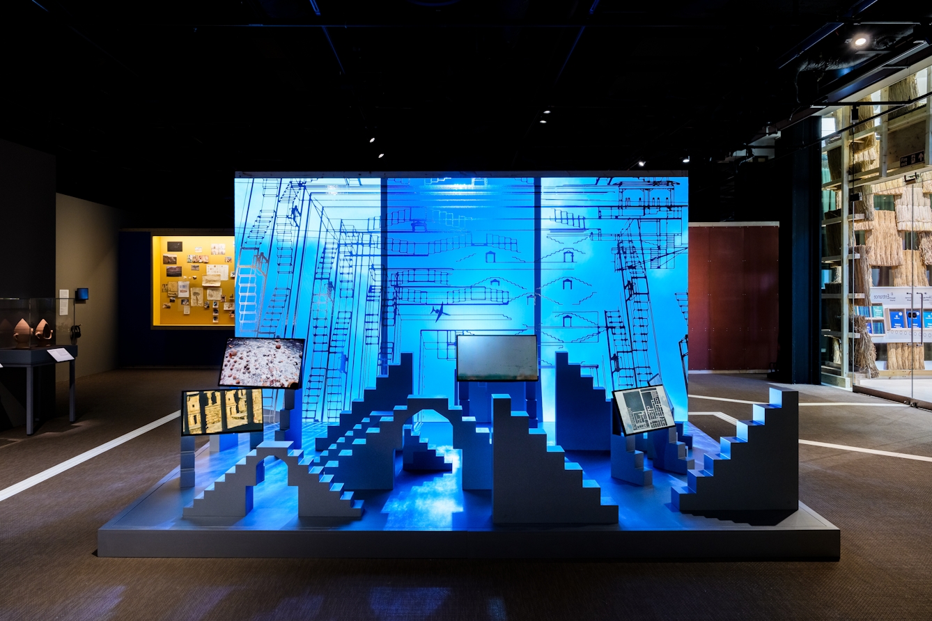An exhibit made up of 3 large frames showing a video of airplanes moving through a cloudy sky, in front of which are a series of stepped and arched sculptures and more video screens.