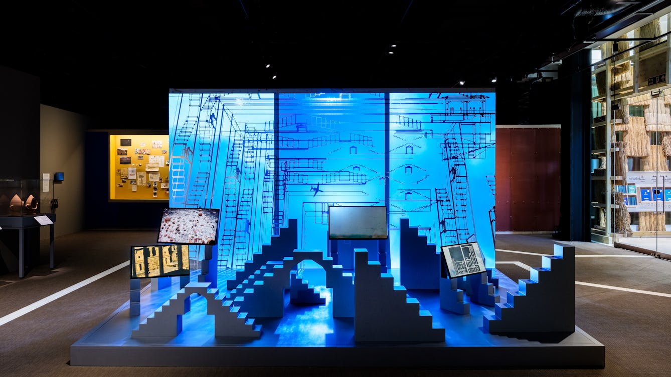 An exhibit made up of 3 large frames showing a video of airplanes moving through a cloudy sky, in front of which are a series of stepped and arched sculptures and more video screens.