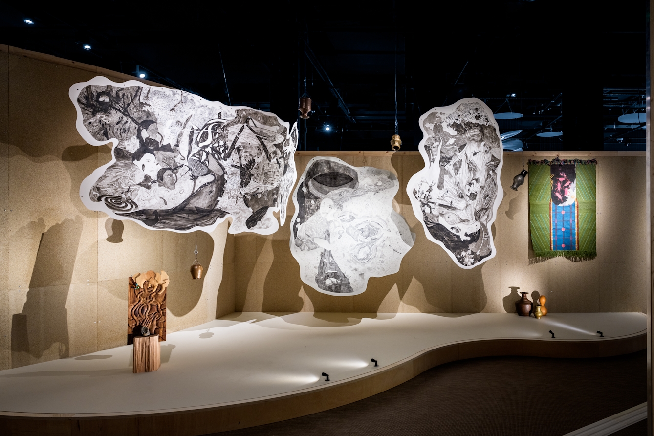 A large artwork installation featuring 3 large handing pen and ink drawings, a wood carving, water vessels and a hanging rug.