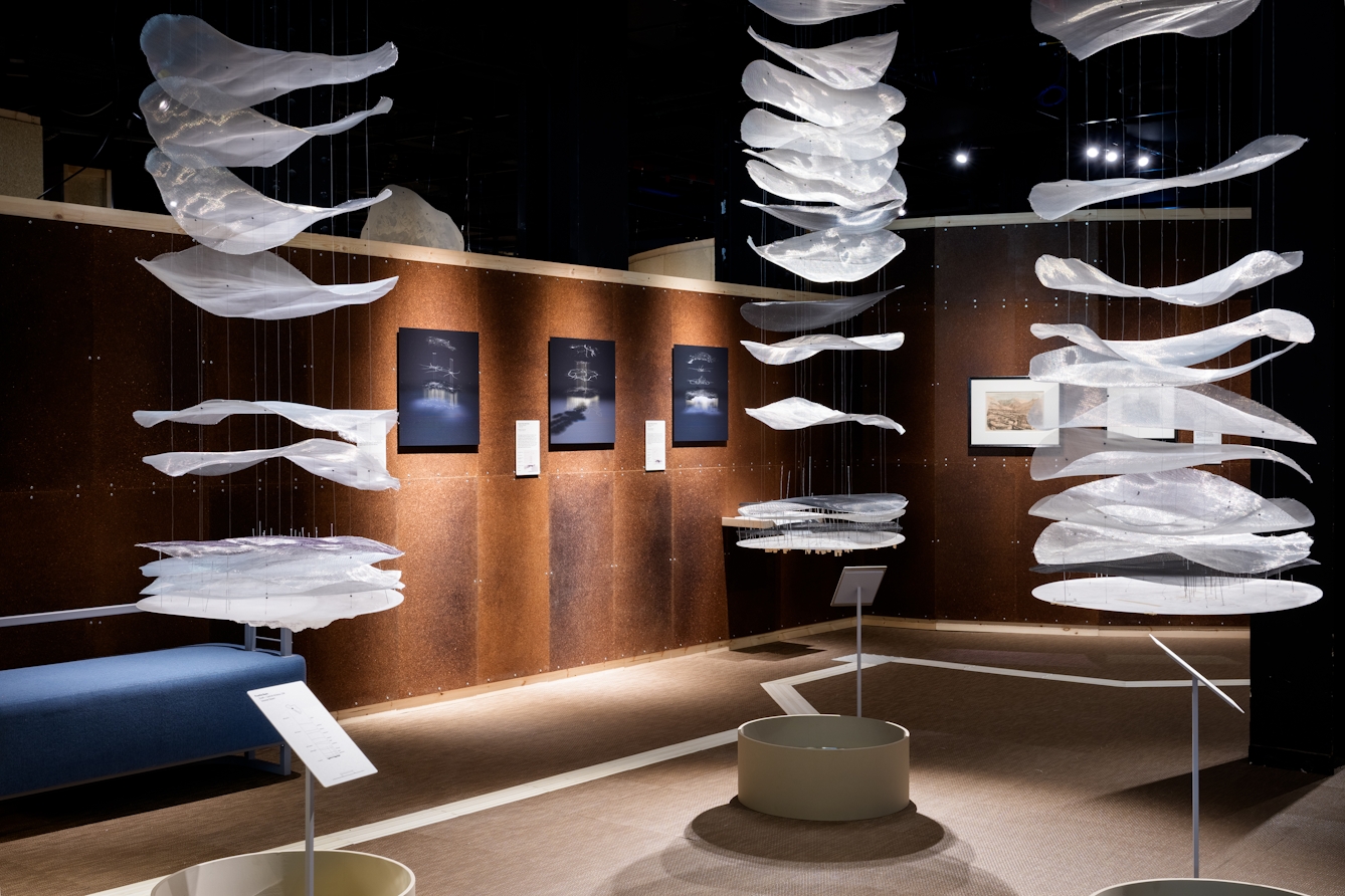 Photograph of gallery installation showing three columns of large white gauze discs, stacked one above the other, hanging from the ceiling. At the base of each is a circular tube containing a mirror.