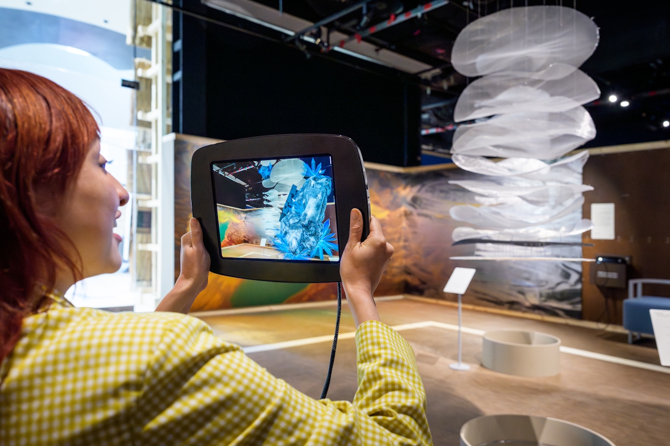 Photograph of a gallery visitor holding up a tablet device to view augmented reality asteroids superimposed on a video feed of the gallery environment from the tablet's camera.