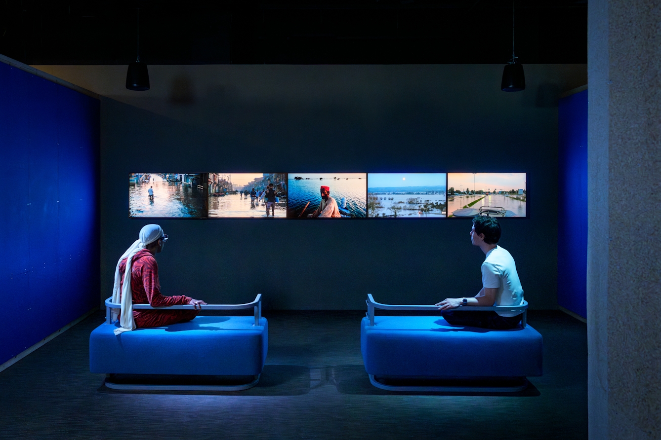 A series of 5 screens mounted in a horizontal line showing films from different parts of the world affected by flooding. Two gallery visitors are sat on benches watching the films.