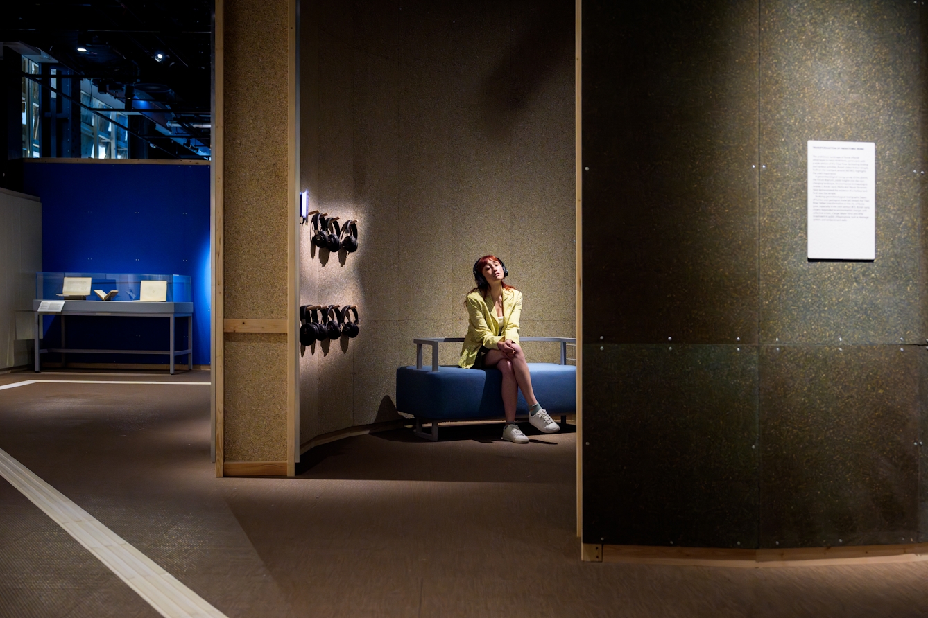 Photograph of a gallery visitor sat in a small circular room, wearing a pair of headphones, eyes closed absorbed in the audio.