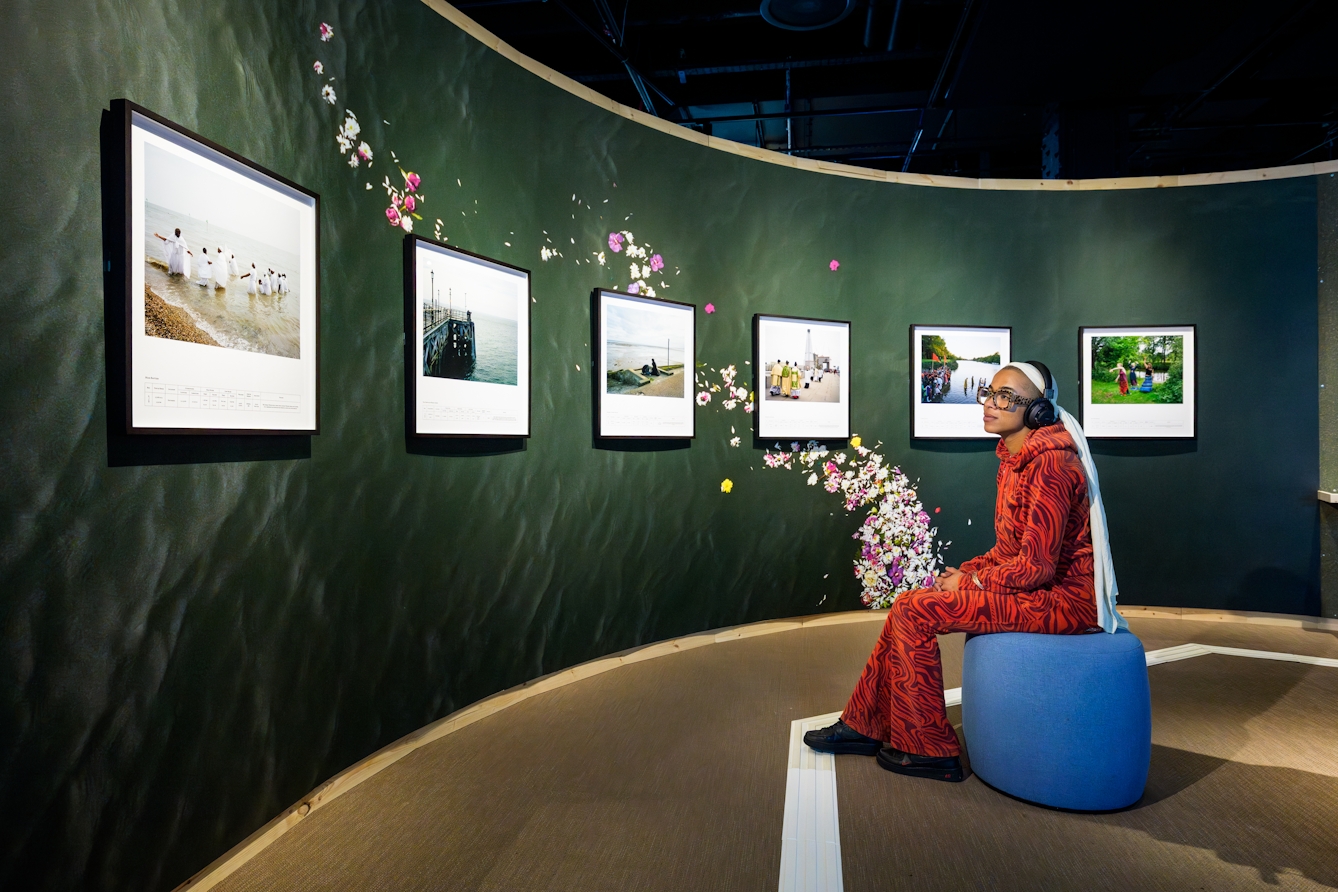 Photograph of a series of six framed colour photographs mounted on a semi-circular wall. A visitor is sat infront of them listing to a commentary on headphones.