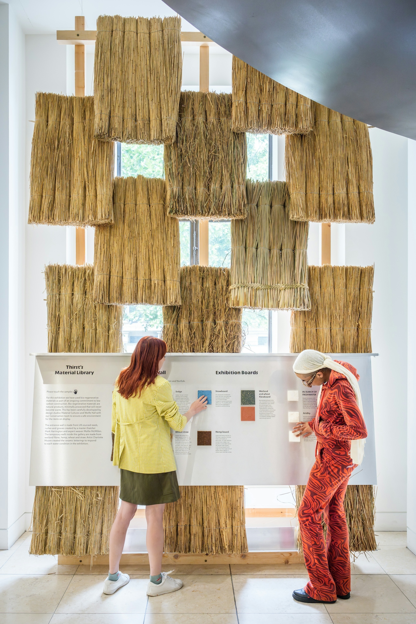 Two gallery visitors investigate a display panel where they can touch and feel some of the materials used in the exhibition. Behind the panel are a grid of thatched tiles.