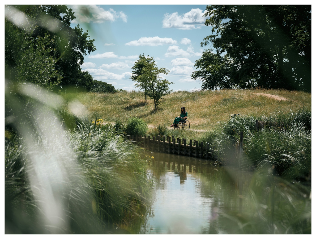 A young woman in the distance, sits in a wheelchair by the side of lake, surrounded by reeds and trees. She is making notes in a small notebook.