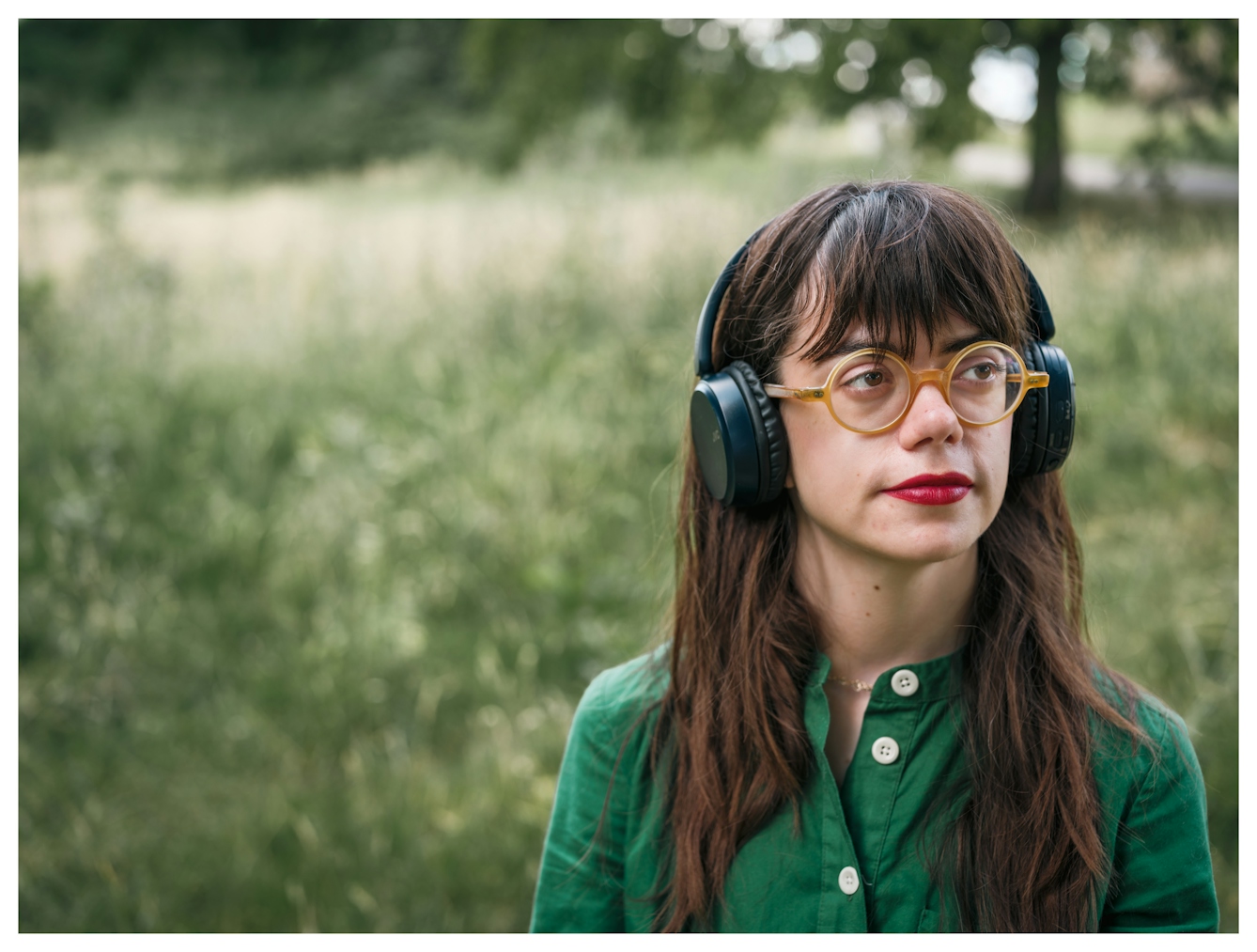 An off centre portrait of Grace Spence Green looking off camera as she listens to music on a pair of over ear headphones. Behind her is a green parkland landscape.