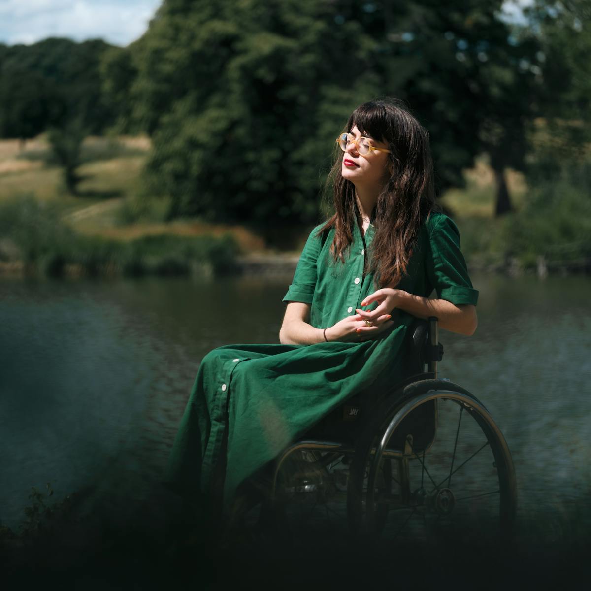 In a park, a young woman in a green dress sits in a wheelchair, eyes closed, bathed in the warm midday sun. Behind her is an expanse of water and trees on the distant hillside.