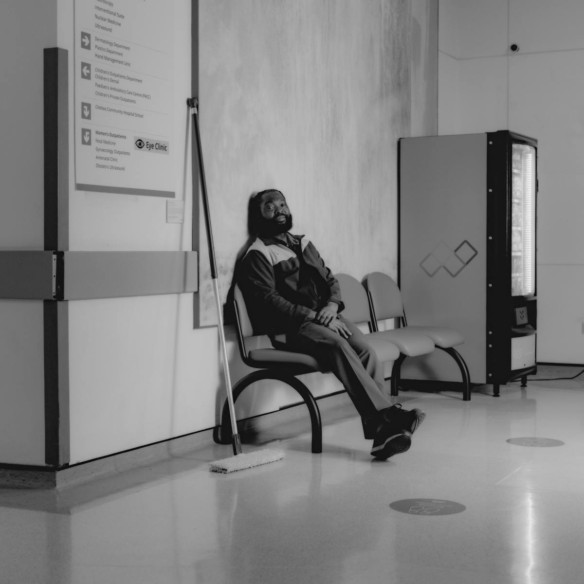 Black and white photograph of a man cleaning areas of a hospital at night. He sits on a waiting area bench, ankles crossed, head resting on the wall behind. He gazes quietly into the camera. Beside him, his mop leans against the wall. He is surrounded by hospital signage and a vending machine.