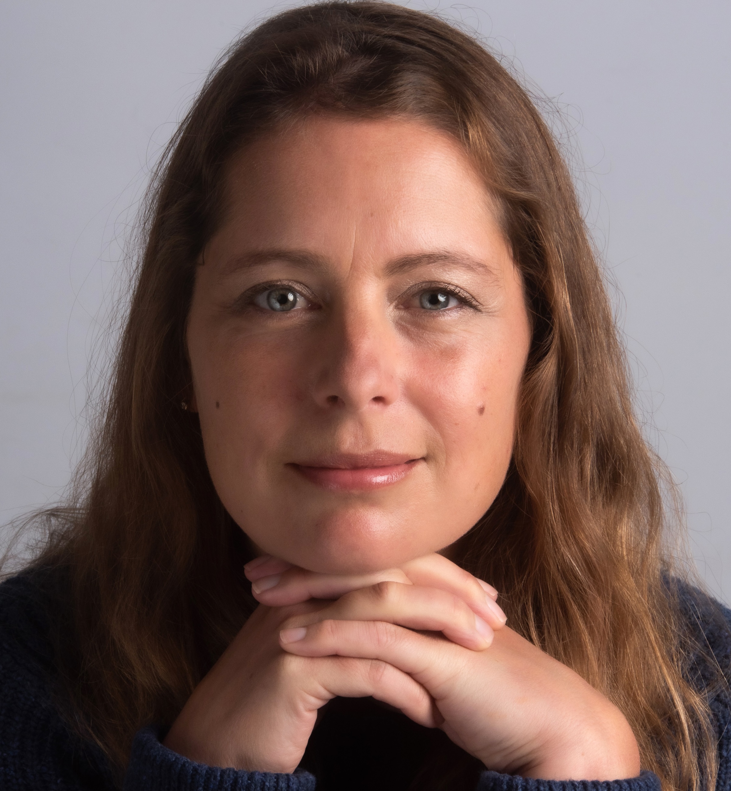 A head shot of a woman with long hair, resting her chin on her hands and smiling directly to camera.