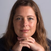 A head shot of a woman with long hair, resting her chin on her hands and smiling directly to camera.