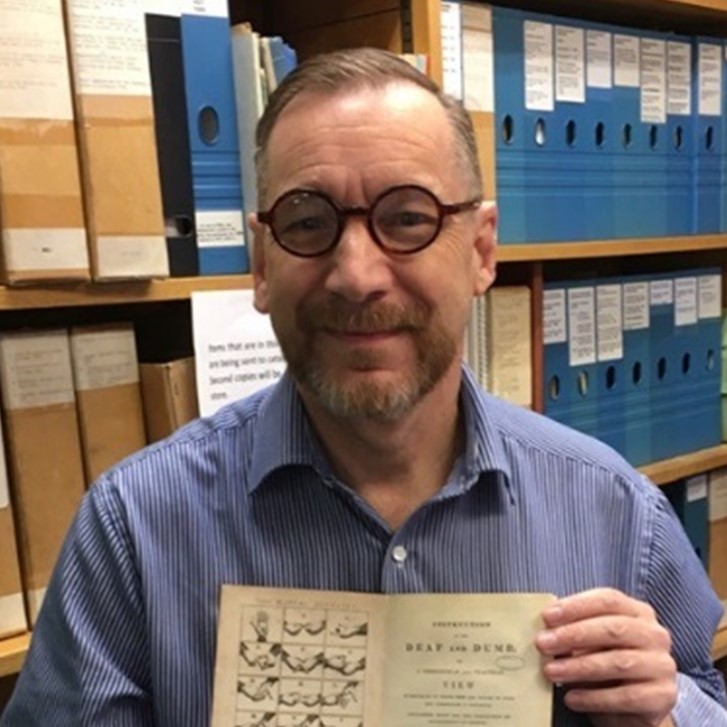 Peter Brown, a man with glasses and a blue shirt, holds a historical book with illustrations of sign language. 