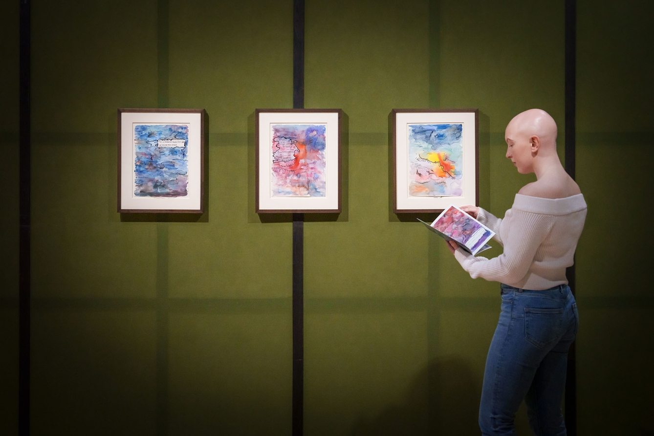 A person stands to the right of a row of three multi-coloured watercolour artworks hanging on the wall in front of them. The person is holding a booklet containing similar artworks