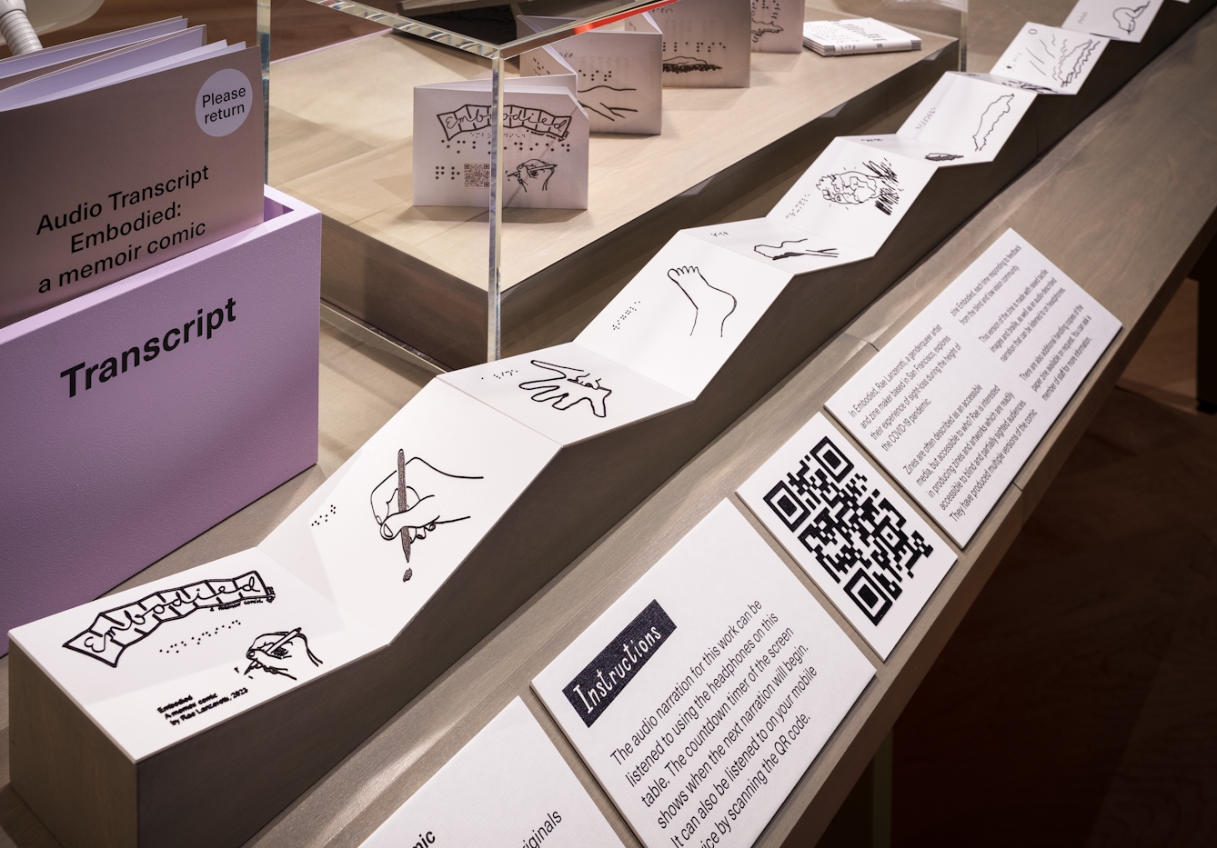 The zine 'Embodied' by Rae Lanzerotti, which consists of a long narrow sheet of paper with multiple folds is shown opened out on a table in front of a display case. Below it are explanatory labels.