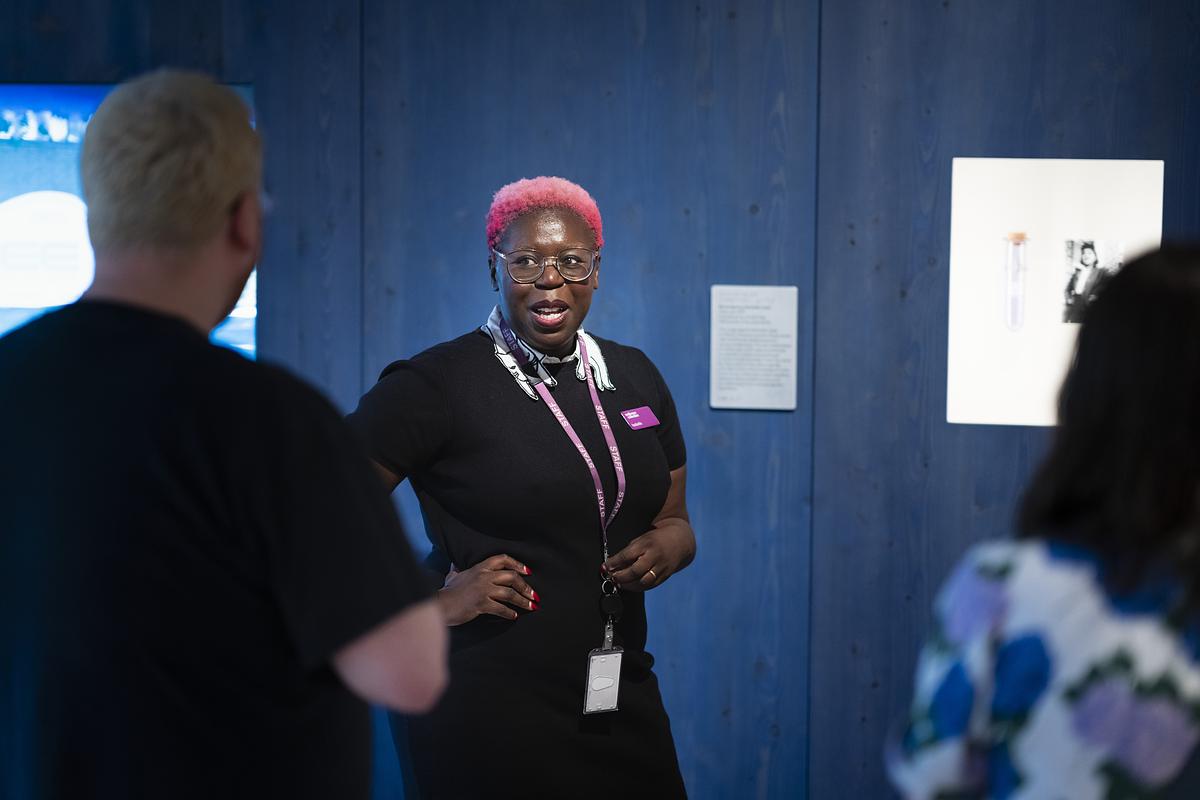 A facilitator with short, bright pink hair delivering a tour to a group of people.