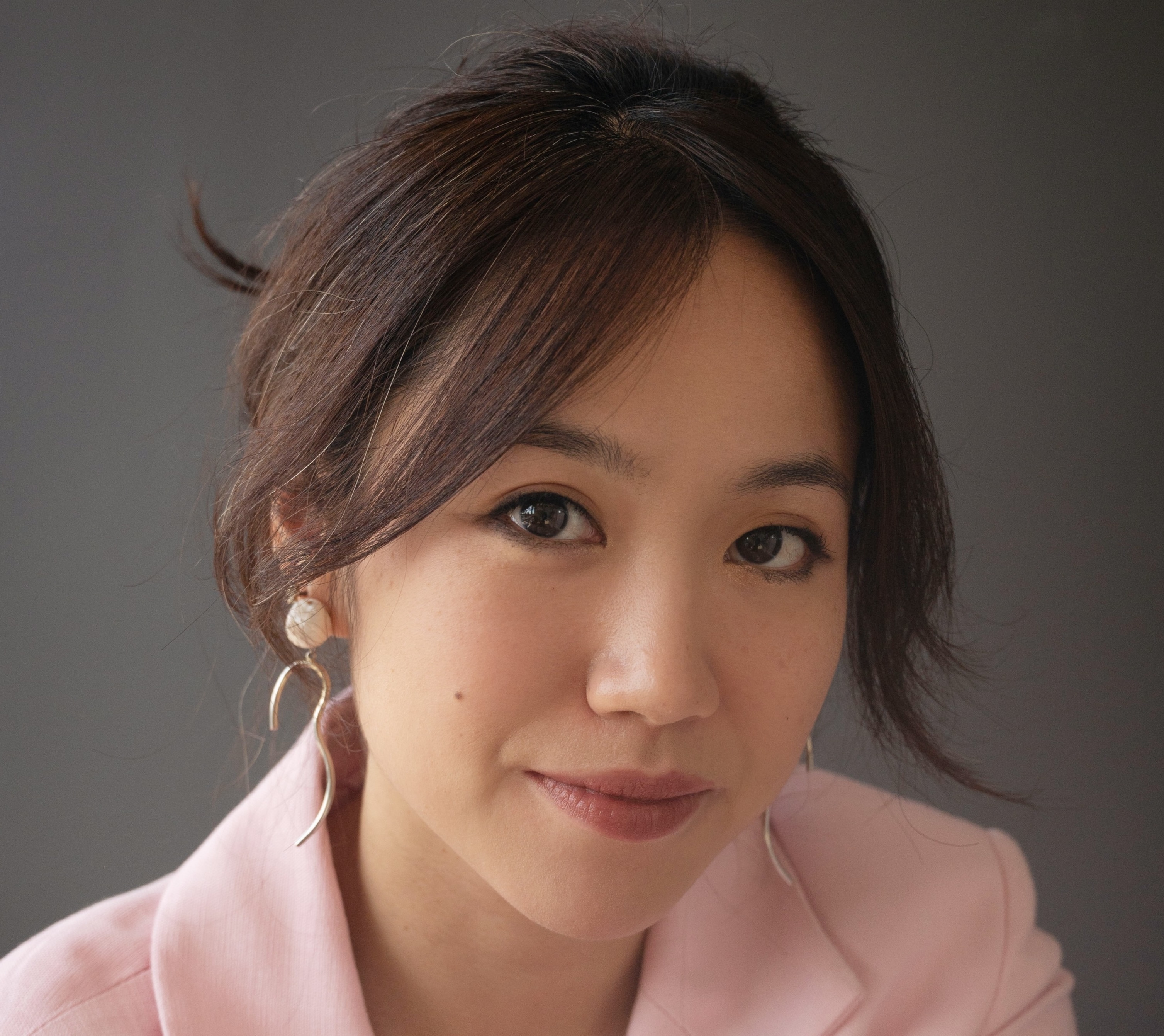 Head and shoulders shot of a woman looking directly to camera, wearing a pink suit jacket with her brunette hair in a loose bun.