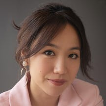 Head and shoulders shot of a woman looking directly to camera, wearing a pink suit jacket with her brunette hair in a loose bun.