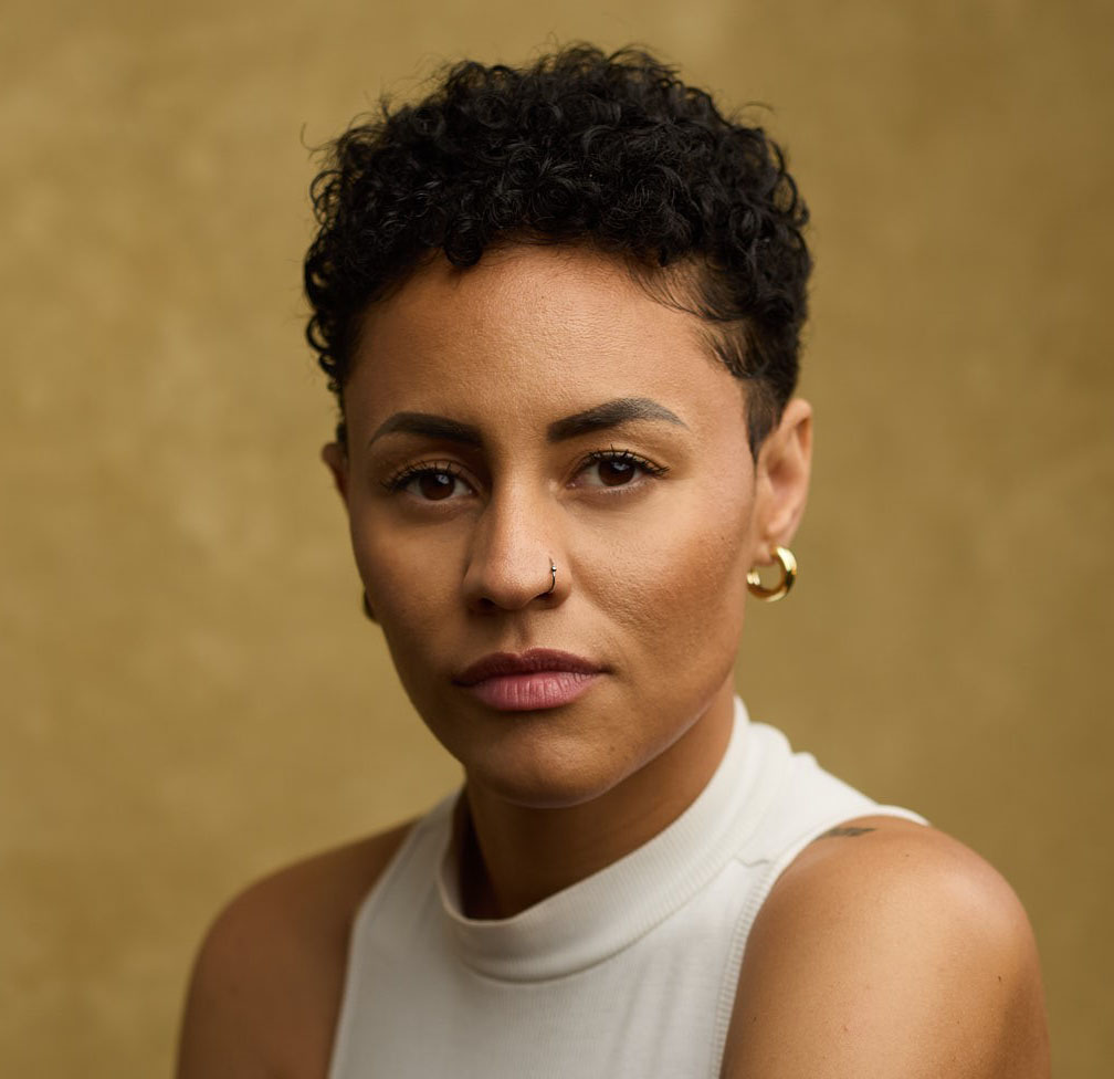 Black and white photograph of the head and shoulders of a young woman with cropped curly hair. She is look directly at the camera.