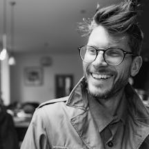 Black and white photograph of the head and shoulders of a bearded man wearing glasses. He is smiling and looking to his right.