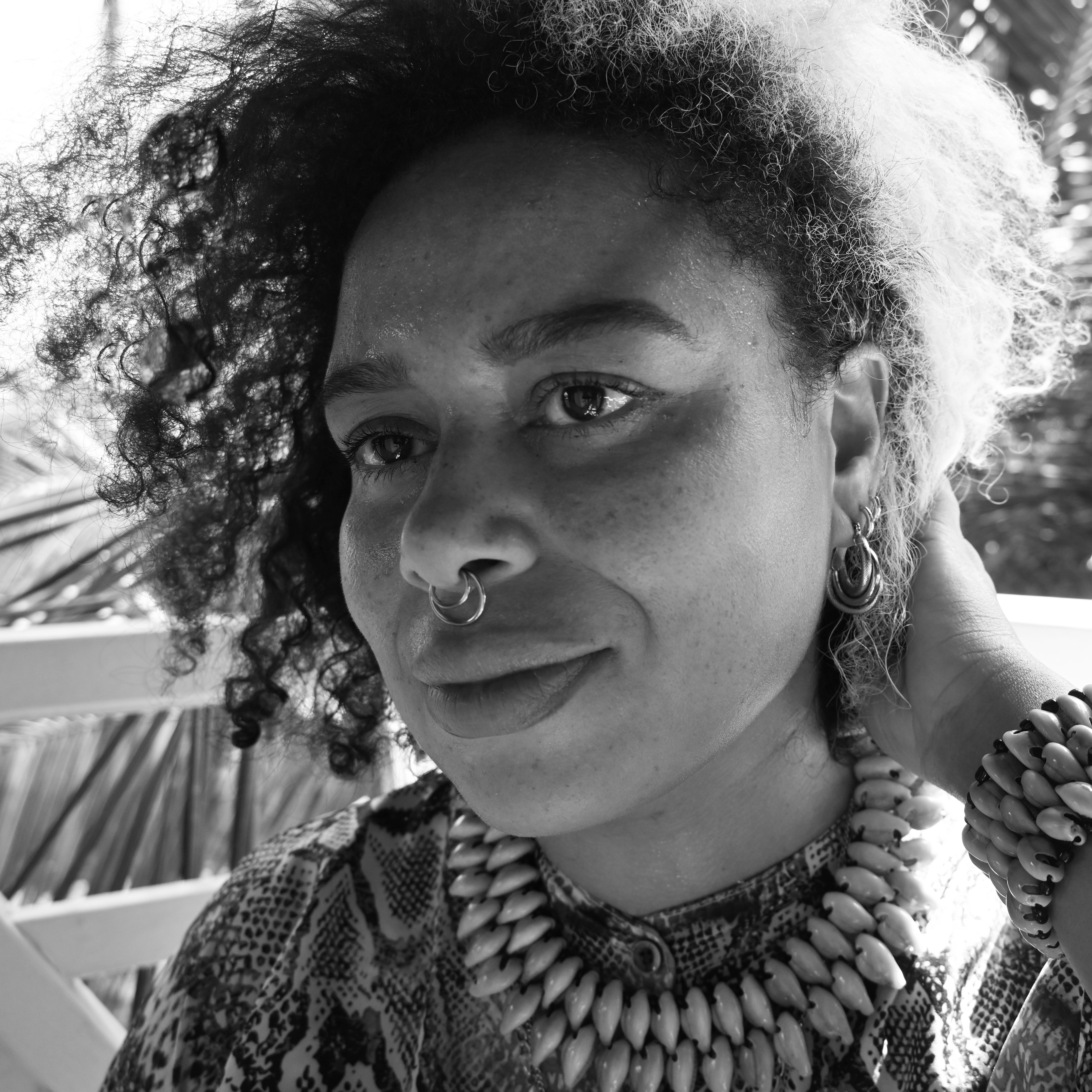 Black and white photograph of the head and shoulders of a young woman with short curly hair, chunky necklace and bracelet, and a nose ring. She is looking to the left-hand side of the camera.