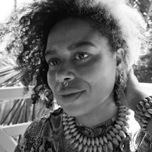 Black and white photograph of the head and shoulders of a young woman with short curly hair, chunky necklace and bracelet, and a nose ring. She is looking to the left-hand side of the camera.