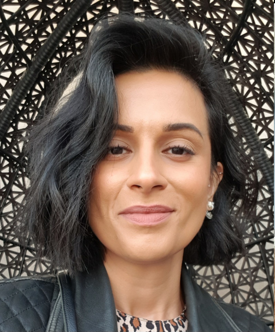 Photograph of the head and shoulders of a woman with short black hair with a blonde streak, leopard print top and leather jacket. She is looking and smiling directly at the camera.