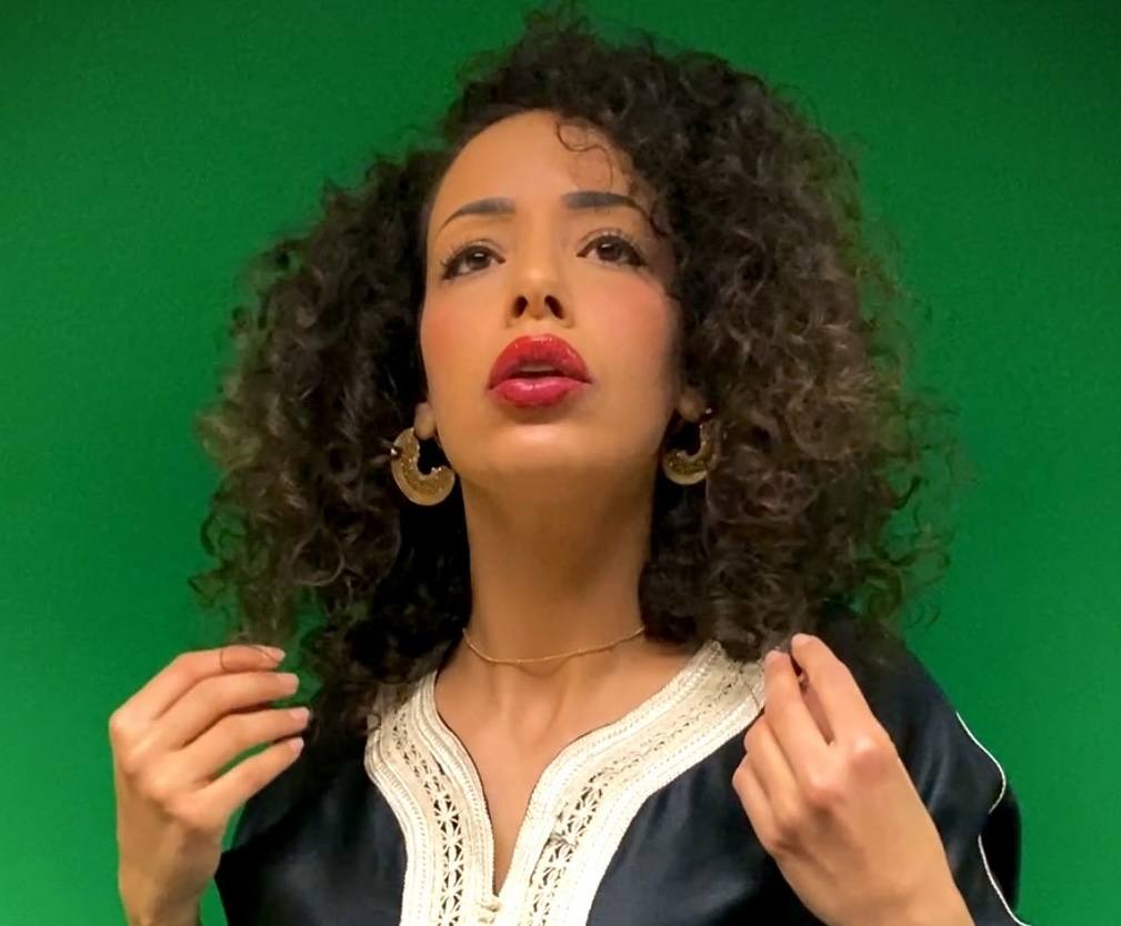 Head and shoulder picture of a woman with shoulder length, curly hair. She is wearing red lipstick, large hoop earrings and is gesturing with her hands and looking above and to the right of the camera.