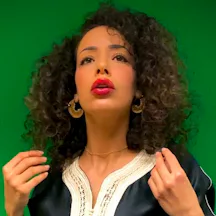 Head and shoulder picture of a woman with shoulder length, curly hair. She is wearing red lipstick, large hoop earrings and is gesturing with her hands and looking above and to the right of the camera.