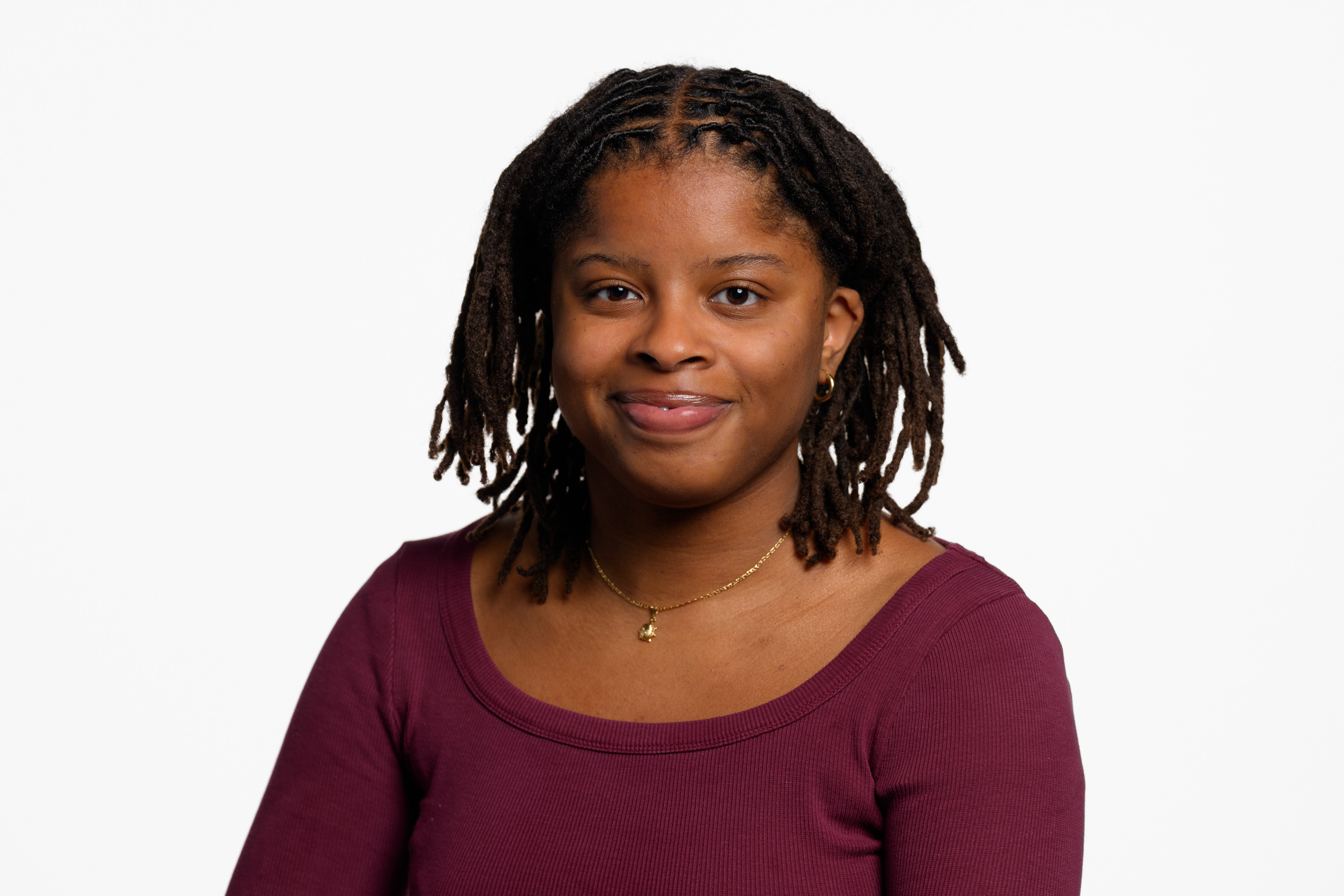 A headshot of Leona Letts smiling at the camera. She has shoulder-length dark hair and is wearing a maroon long-sleeved T-shirt, a gold pendant necklace, and small hoop earrings.