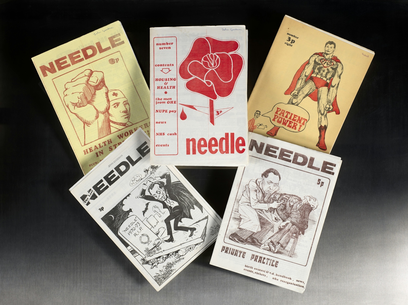 Five Needle magazines are displayed on a dark surface in a scattered arrangement. Each cover features distinct graphics and titles related to healthcare themes, such as a raised fist, a stylised rose, a superhero with a "NHS" emblem, a cartoon of a vampire next to a gravestone reading "'Needle' 1970-73 R.I.P", and a doctor examining a patient. The magazines use bold illustrations and a mix of red, black, and brown tones, highlighting their activist and satirical content.