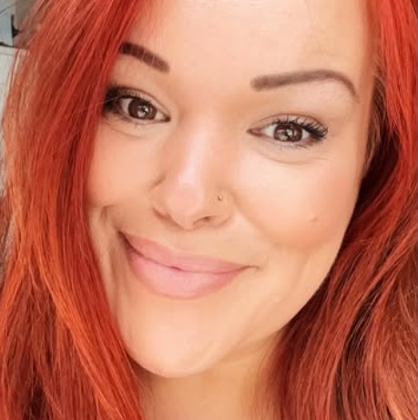 Close-up headshot of a woman with long red hair, smiling to camera.