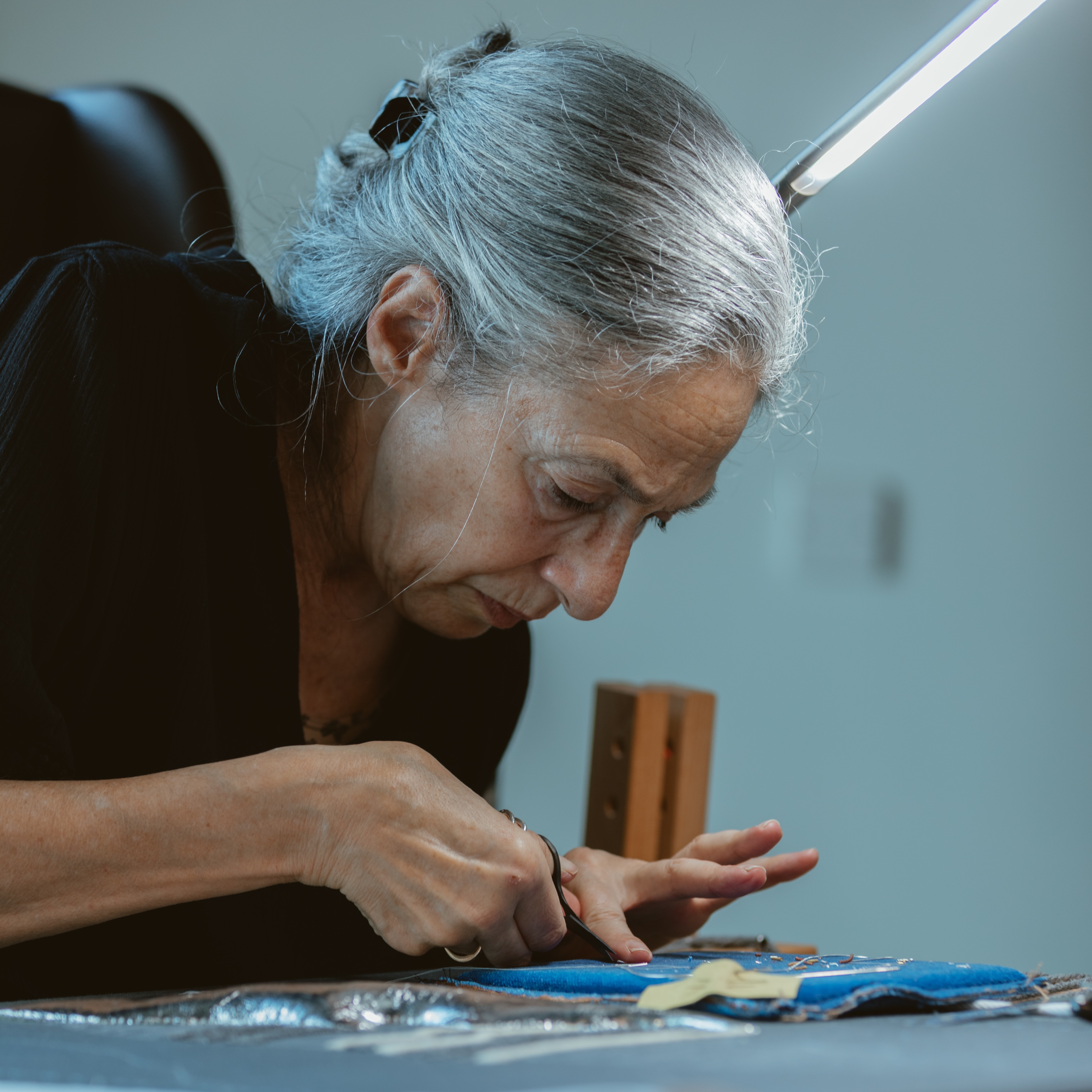 A person with grey hair tied back, wearing a black top, is focused on cutting fabric with scissors at a worktable. Various sewing materials and tools are laid out on the table, and a bright light illuminates the workspace.