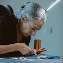 A person with grey hair tied back, wearing a black top, is focused on cutting fabric with scissors at a worktable. Various sewing materials and tools are laid out on the table, and a bright light illuminates the workspace.