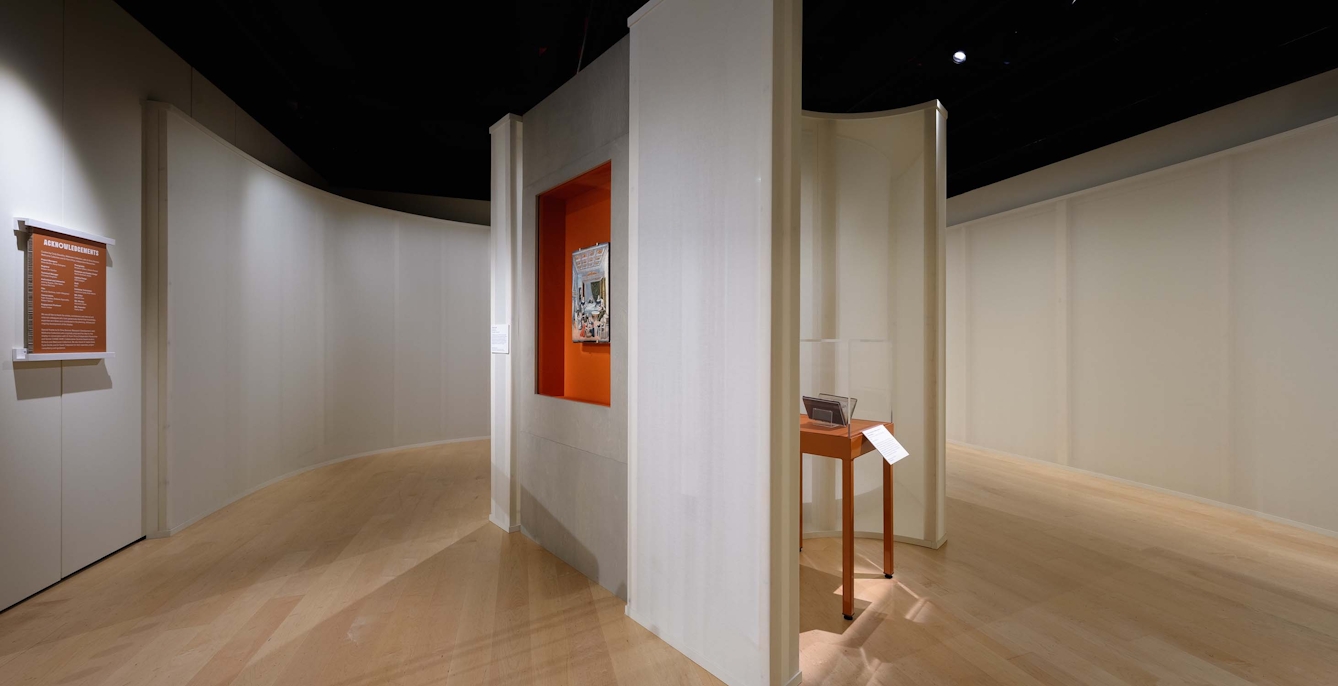 A museum gallery space showing a room divided by a wall with two artworks on opposite walls in one half and a display case against a wall in the other half.