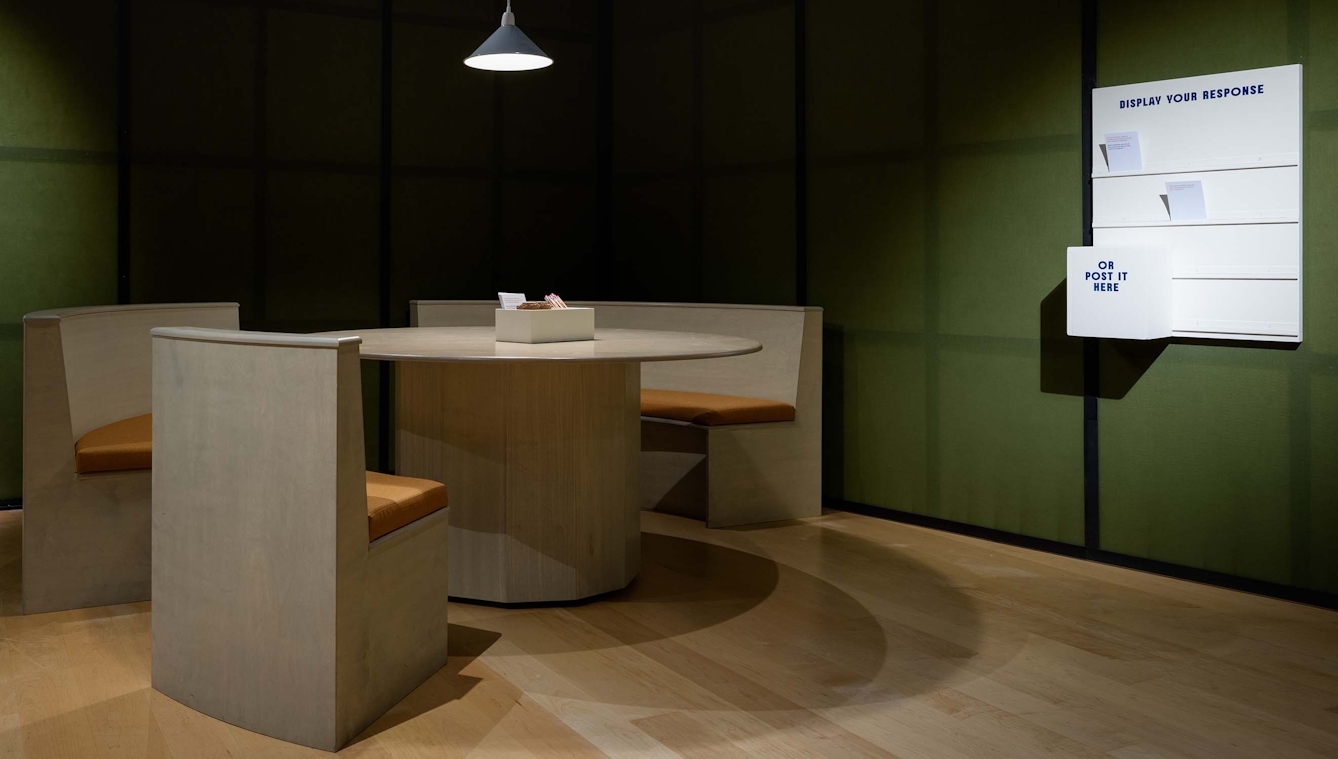 A table and three seats in the corner of an exhibition gallery.