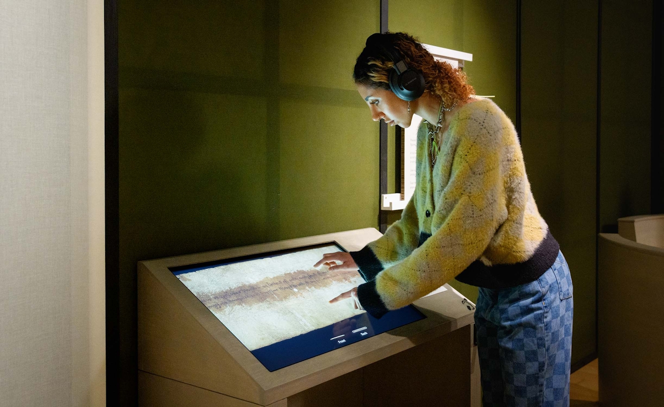 A woman wearing headphones is touching an interactive display screen in an exhibition gallery.
