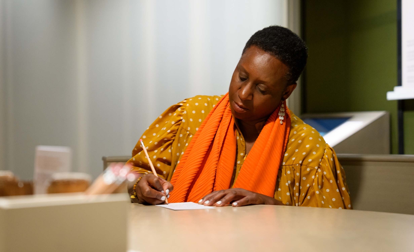 A woman sat at a table is writing on a piece of paper in front of her.