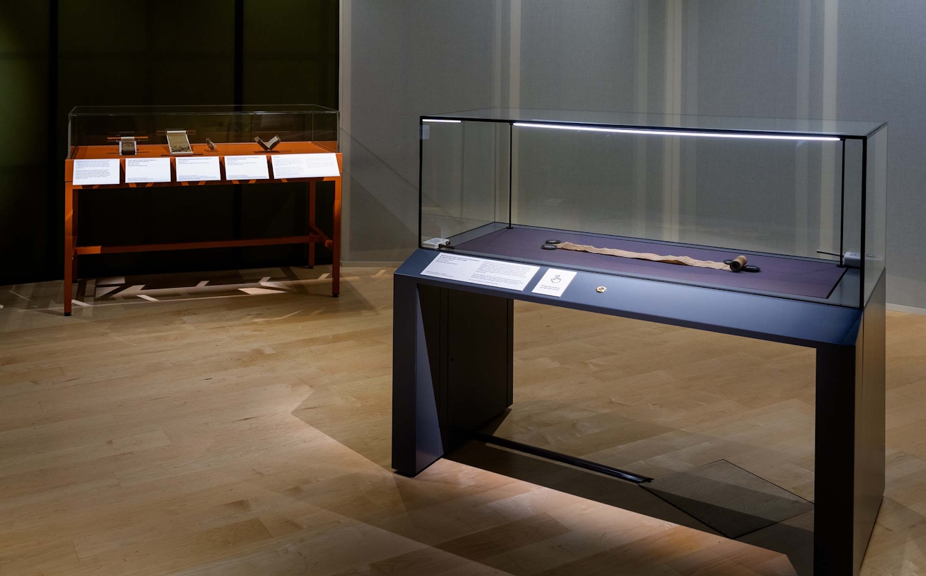 A glass display case in an exhibition containing a medieval parchment scroll, partially unrolled. Against the back wall is another display case with more objects.