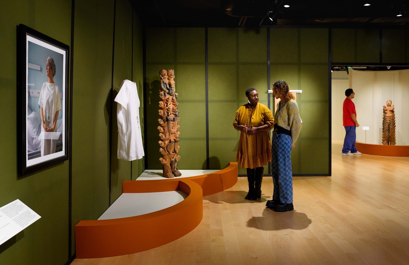 Two people stand infront of a long white garment on display in an exhibition, They are talking to one another.