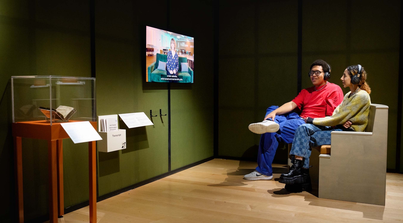 Two people are seated infront of a video screen mounted on the wall in an exhibition gallery. They are wearing headphones and watching a video.
