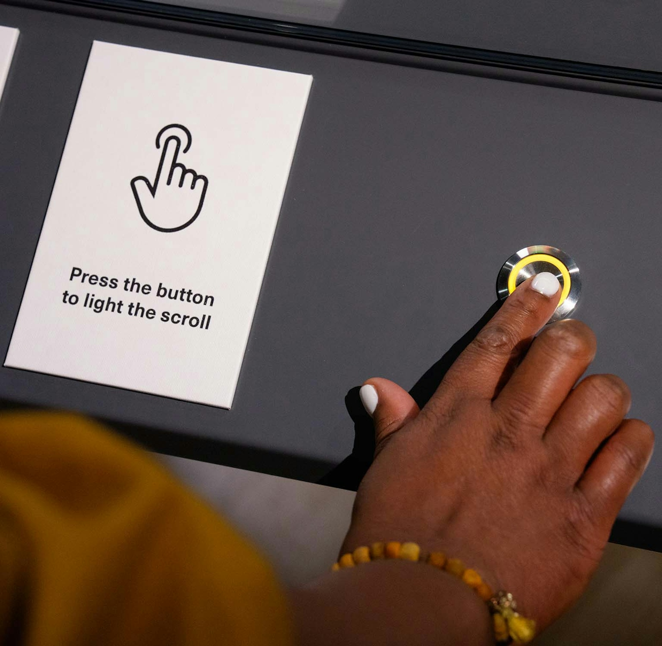 A hand pushes a button mounted in a table with one finger. Next to the but is a sign with a drawing of a hand pushing a button and the text: "Press the button to light the scroll".