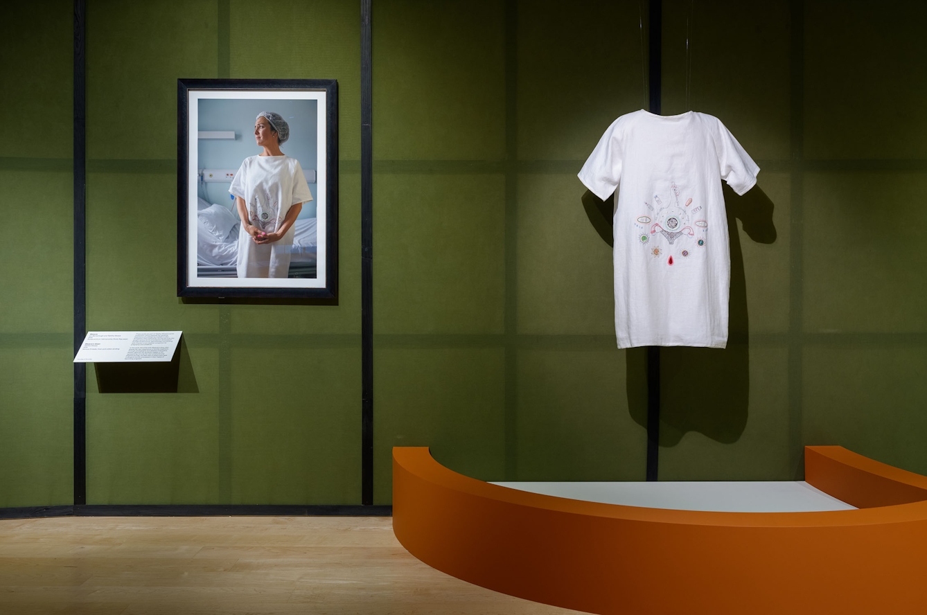 A white garment with an embroidered pattern on the front hands next to a framed photograph of a woman wearing the gown in an exhibition gallery. The embroidery is of a womb and motifs relating to IVF treatment.
