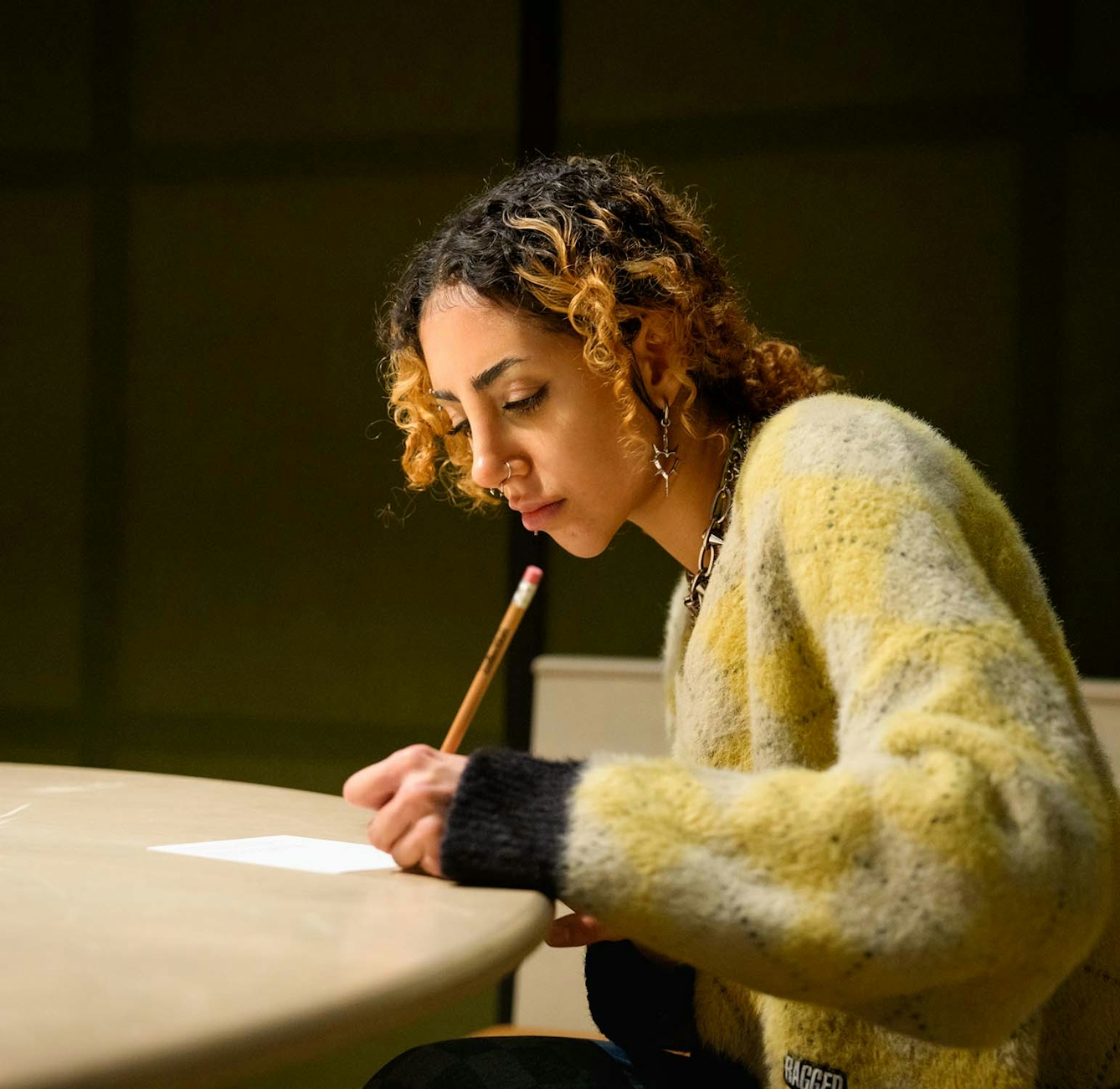 A young woman sits at a desk with a pencil in her hand. She is writing on a small rectangular card of white plane paper.