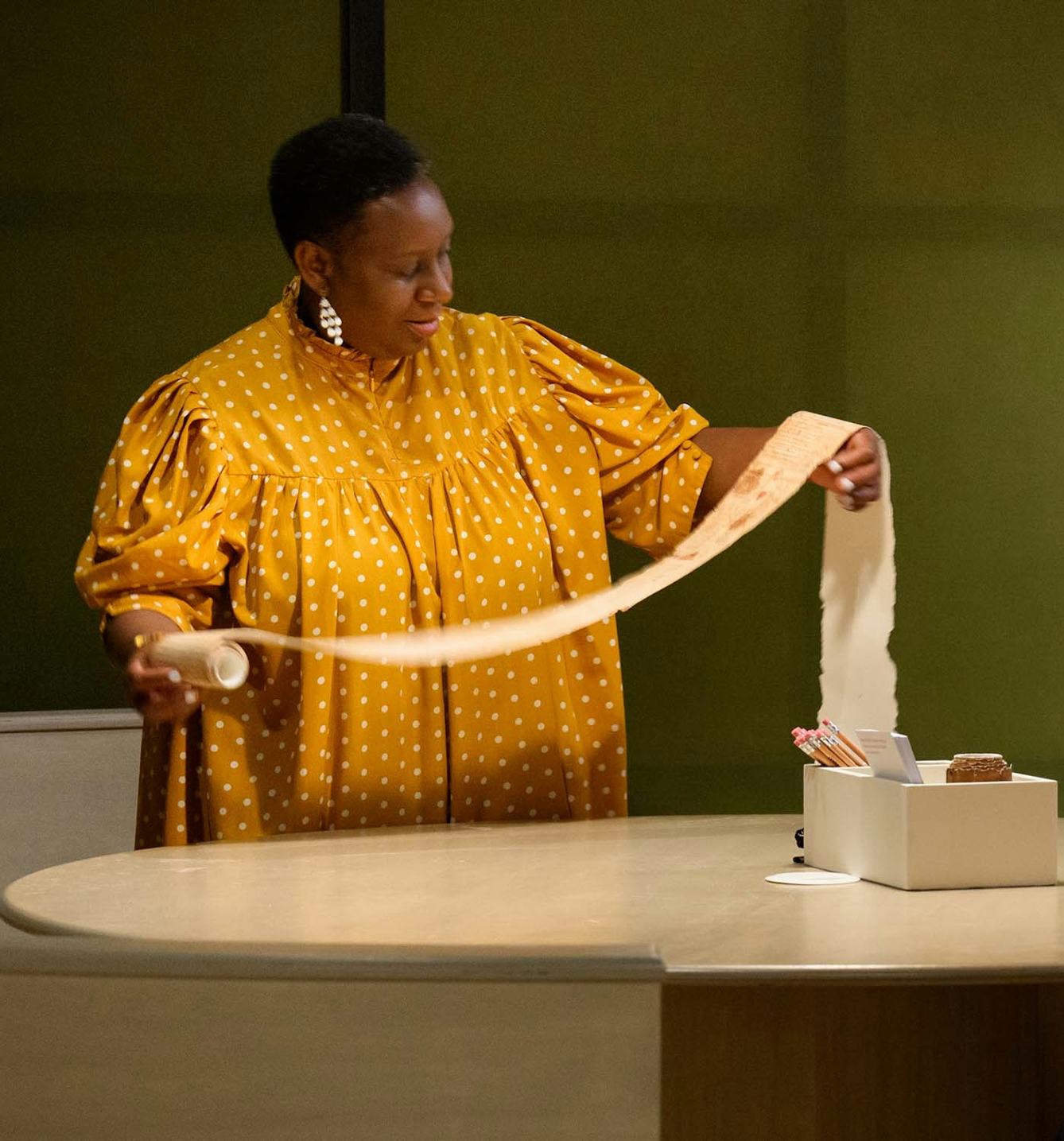A woman unrolls a long scroll of paper in front of a table.