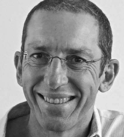 Black and white photograph of the head of a man wearing rimless glasses. He is smiling and looking directly at the viewer.