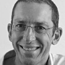 Black and white photograph of the head of a man wearing rimless glasses. He is smiling and looking directly at the viewer.