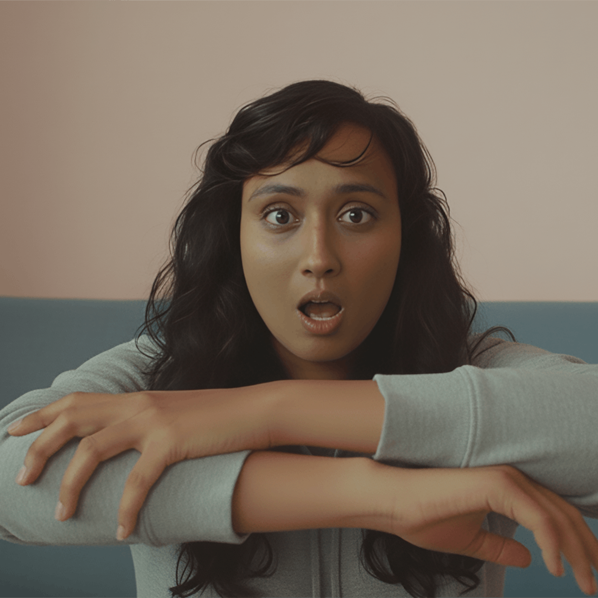 A digital illustration of Sabrina as an adult, seated indoors on a muted blue sofa, saying “brown” in British Sign Language. She has long brown hair and her arms are crossed horizontally in front of the chest, with her hands positioned to form a clear sign.