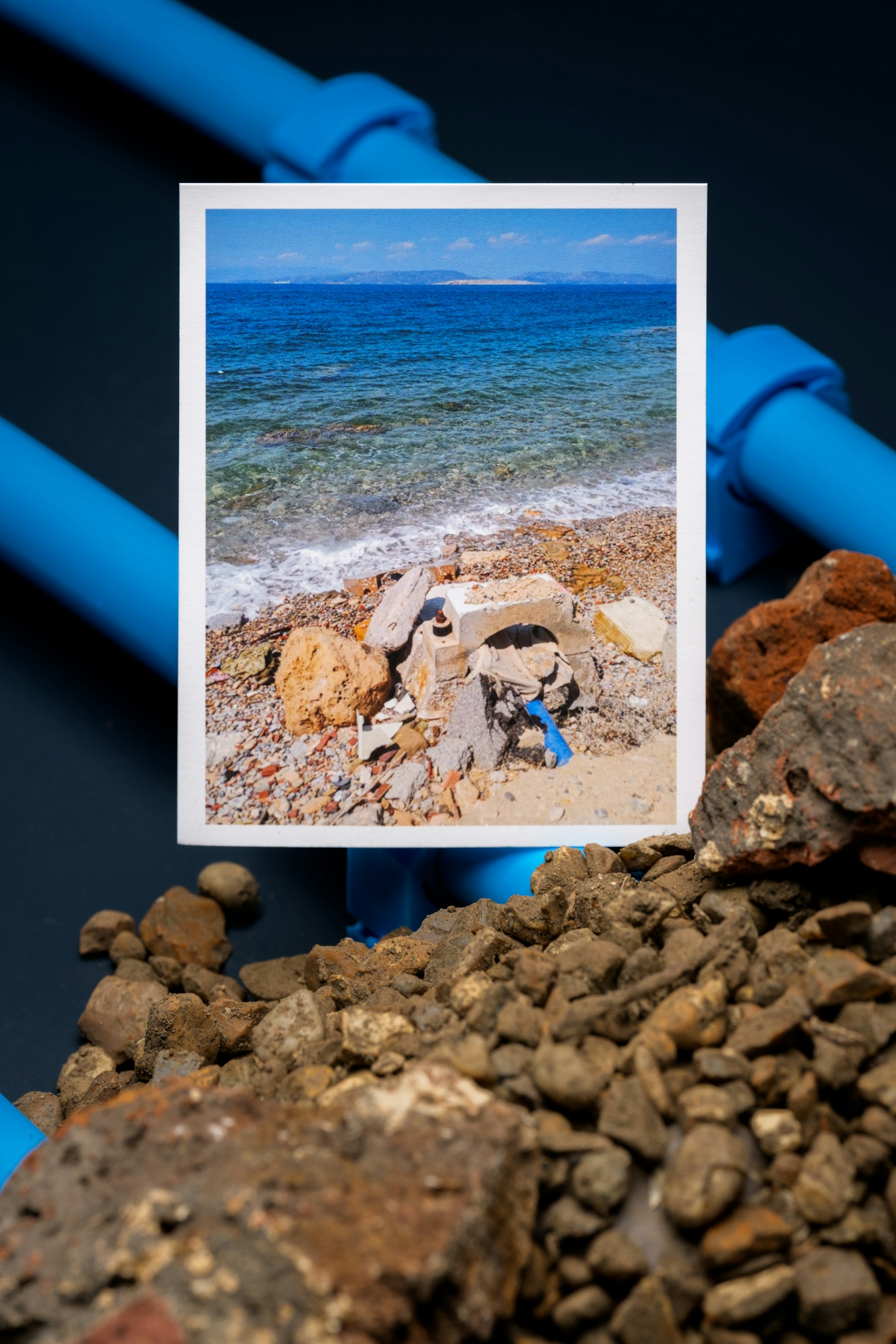 A photograph of a stone coastline with a blue water pipe buried under rocks. The photograph is positioned on a background of blue water pipes and stones.